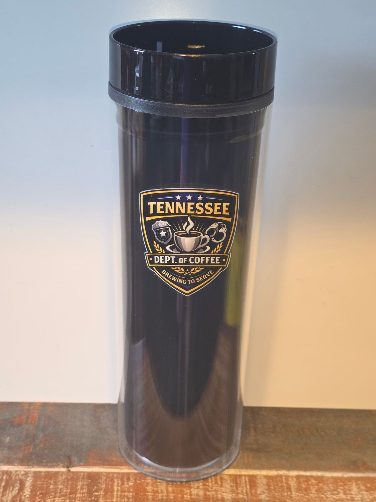 A tall, clear double-wall insulated tumbler with a black lid, featuring a Tennessee 'Dept. of Coffee' shield logo printed on the front, set on a wooden surface against a plain light background.